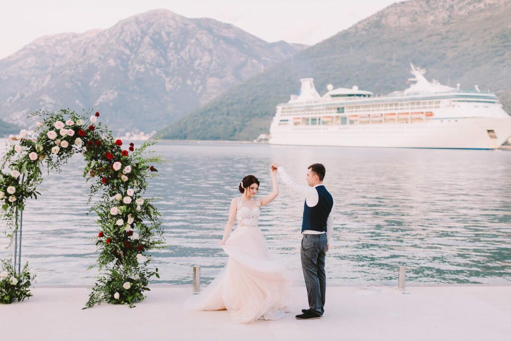 Wedding couple by floral arch on the waterfront at Adriatica, Kamenari, Boka Bay, Montenegro. Caption: Portraits with Boka Bay behind you.